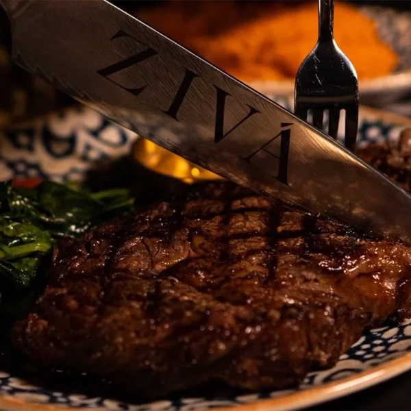 Close-up of a grilled steak served with sautéed vegetables, with a knife engraved ZIVÁ resting on top.