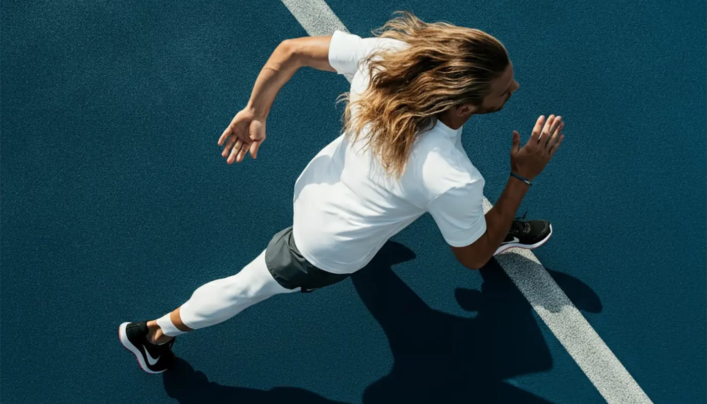 Athlete with long hair sprinting on a blue running track, captured from above in mid-stride.